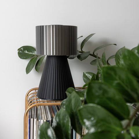 Lustra Plissé table lamp in Smoke Black next to green foliage in daylight