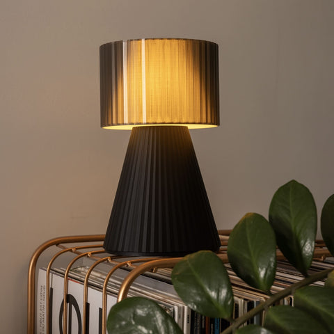 Lustra Plissé Smoke Black lamp glowing warmly on side table with plant