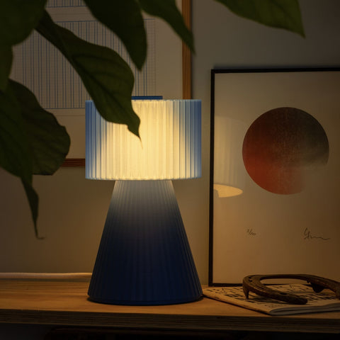 Lustra Plissé Ocean Blue lamp glowing softly behind indoor plant in cozy room
