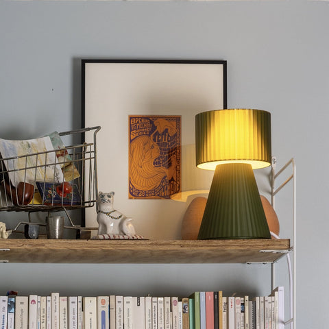 Lustra Plissé Forest Green lamp glowing warmly on shelf beside framed art