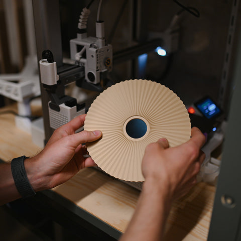 Person holding a beige circular object with a blue center in front of a 3D printer.